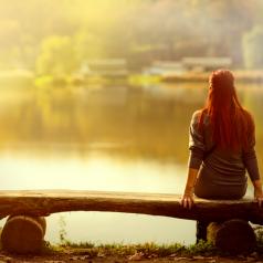 Rear view of person with long red hair sitting on bench at sunset looking over lake