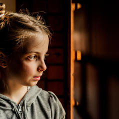 young child with hair up in pigtails looks off to the side in dimly lit room with a serious expression