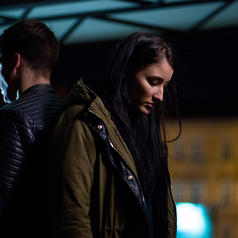 Couple dressed in light jackets stand outside at night at distance from blurred lit building, facing away from each other, looking down
