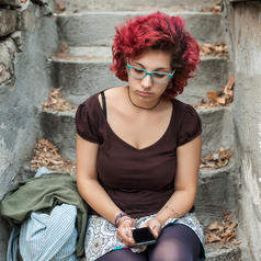 Person with short curly red hair, blue glasses, wearing tights and skirt sits on leaf-strewn stairway and reads on phone with somber expression