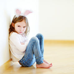 Very angry child with long hair and rabbit ear costume sits on floor with arms crossed, glaring