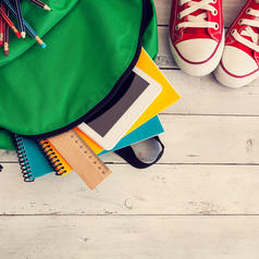 Green backpack lies open, school supplies sticking out of the top, on wood floor next to red sneakers