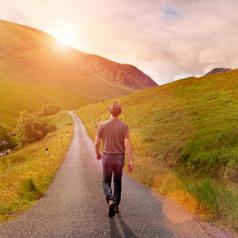Rear view of person in casual clothes and hat walking alone on a narrow country road through a green valley surrounded by mountains, heading toward the rising sun. Warm sunlight with lens flare. Taking decisions and choosing a direction for new beginnings, following the way forward, not looking back.