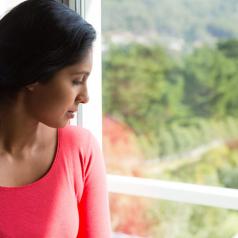 Adult wearing pink top with long hair pulled back looks out window into fields and trees, thoughtful expression on face