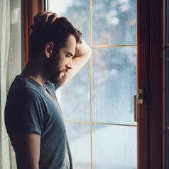 Young adult with short hair, facial stubble, leaning on one arm looking out windowed door during rain