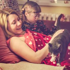 Parent and young child with glasses, barrettes, braids sit on daybed and pet cat