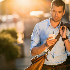 Person walking down sunny street looks at phone, holding coat over shoulder with one hand