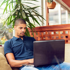 Person with shaved head wearing polo shirt sits in bright cafe and uses laptop computer with serious expression