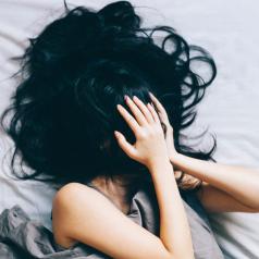 Person with long black hair lies in bed, hands and hair over face