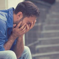 Person with short hair in work shirt crying on stone steps outside, covering face in hands