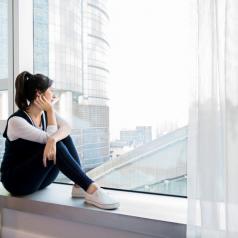 Young adult with ponytail in casual comfortable clothes sits on windowsill, leaning cheek on hand and looking out