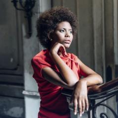 Young adult in red blouse with natural hair stands leaning on banister, lost in thought, with serious expression