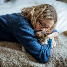 Person in blue sweater with long wavy blonde hair and glasses lies on bed with somber expression holding cat and cuddling 