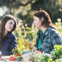 Mother and daughter sitting at a table outdoors on an organic farm, conversing on a bright, sunny day. On the table is fresh produce, including purple cabbage, radishes, leafy green vegetables and a basket of eggs.