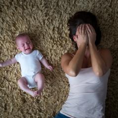 Stressed mother lies on back on carpet next to fussing baby, covering face with hands in distress