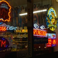 Photo shows neon signs advertising various alcoholic beverages in window of street storefront at night
