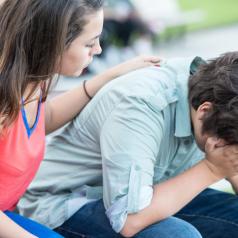 Young teen leans forward with face in hands while friend places comforting hand on back