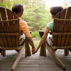 Rear view photo of older couple sitting on deck in Airondack chairs holding hands