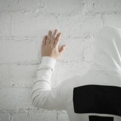 Photo shows back of young teen wearing a hoodie standing with palm on whitewashed wall