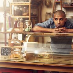 Non-white server waits behind counter of bakery/cafe for customers, resting chin on hands
