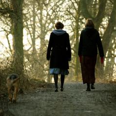 Rear view photo of two people in long skirts and cold-weather wear walking dog along forest path