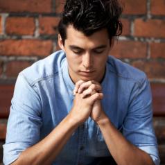 Young adult sits on bench outside brick wall, praying with eyes closed