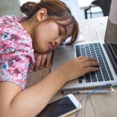 A teen girl in a pink shirt rests her head in front of a laptop. 