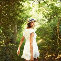 Young adult wearing dress and matching summer hat stands on forest path looking back and smiling