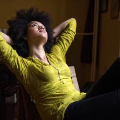Young person with natural hair wearing green blouse leans back in chair, thoughtfully looking up 