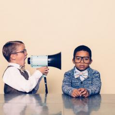 Two boys in bow ties and glasses sit at a table. One is yelling through a megaphone.