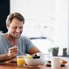Person wearing t-shirt with short hair and facial hair sits at kitchen table eating breakfast