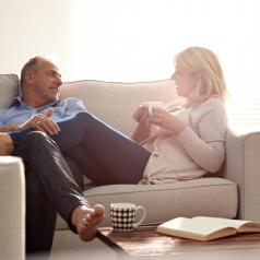 Mature couple relaxing together on sofa hold coffee and smile at each other in living room 