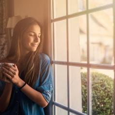 Person with long hair looks out window of small house, smiling, holding coffee, wearing casual clothes