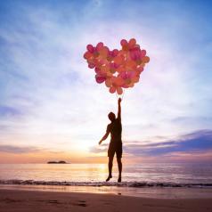 Person at beach holds heart-shaped bunch of balloons and rises slightly off ground