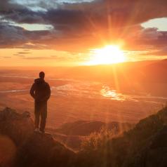Person stands on a ledge of a mountain, enjoying the sunset over a river valley 