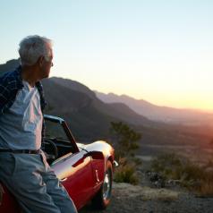 A senior man leans against his car as the sun rises.