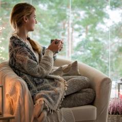 Person with long hair in braid sits looking out window holding hot drink wrapped in sweater
