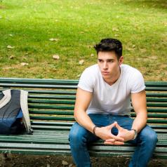 Young student with short hair sits on bench, looking lonely, backpack nearby
