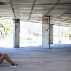 Teenager with hair in ponytail sits in stone building looking down alone