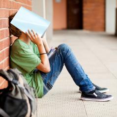Teenager leans against brick wall with notebook open over face, backpack nearby