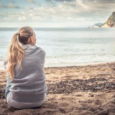 Thoughtful young adult with long hair in ponytail sits on beach and looks out over water