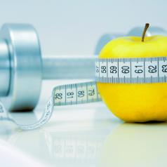 Close-up of an apple with a measuring tape around it and chrome dumbbells at the background. 