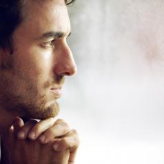 Young adult with short hair and facial hair, hands clasped under chin, looks into distance thoughtfully
