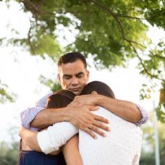 Hispanic parent hugs two teenage children outside under trees 