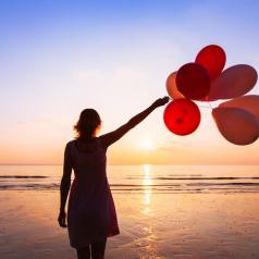 Person looks out to sea, raising arm with bunch of balloons to sky