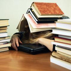 A young woman is sleeping on a laptop with a heap of books on her head.