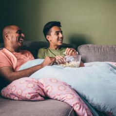 Father and teenager sit on sofa with popcorn in living room watching something on TV together