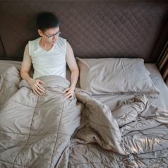 Young Asian adult with short hair and glasses sits up in bed under duvet cover looking downcast