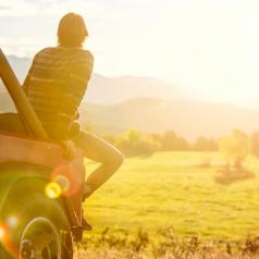 Rear view photo of young adult with short hair sitting on back of Jeep looking out at mountains and field