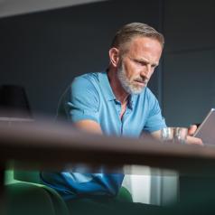 Stressed-looking mature adult with gray hair and short beard looks serious at table while reading on tablet